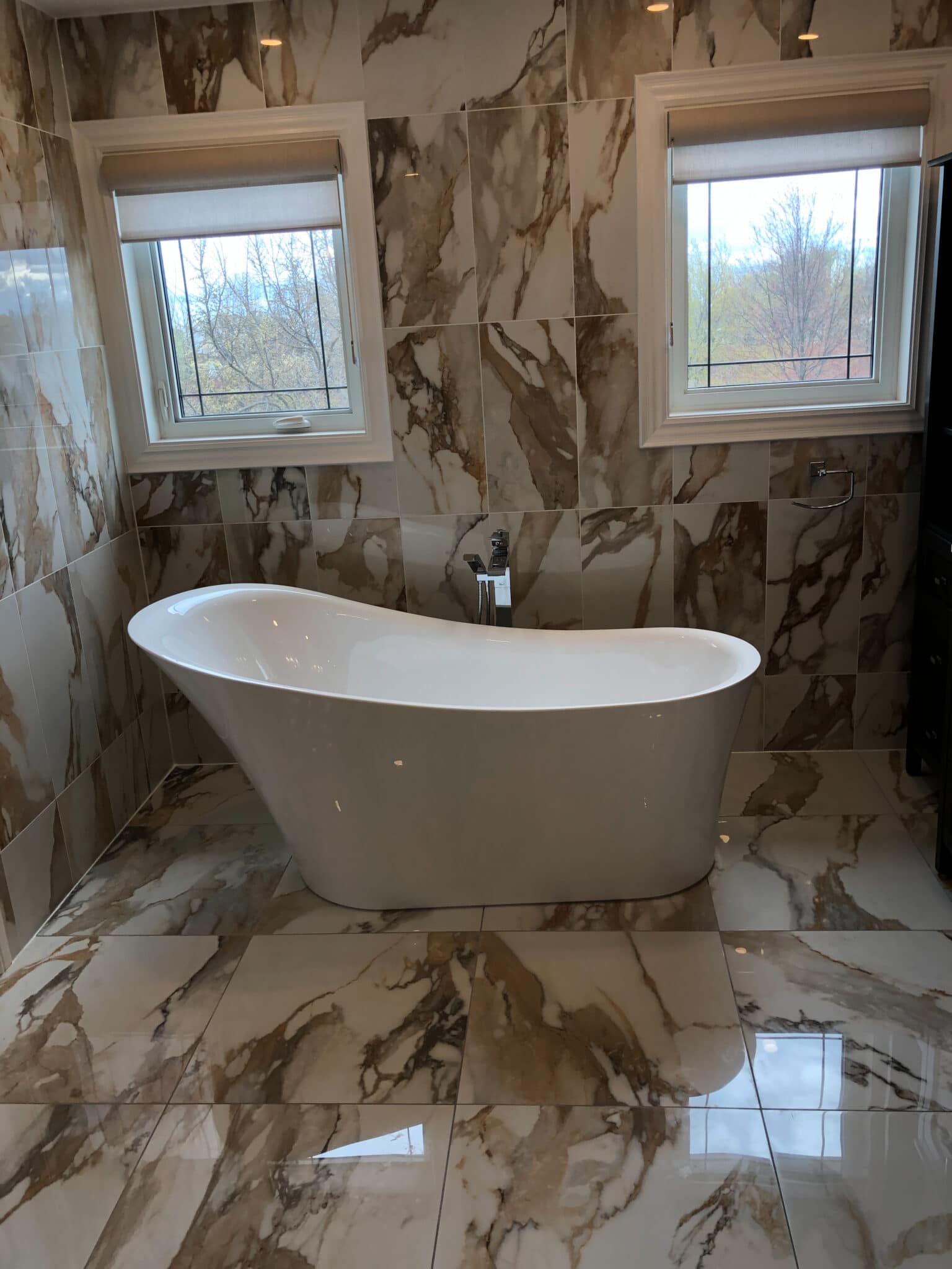 Modern bathroom featuring a freestanding white bathtub, large marble tiles with a brown and beige pattern, and two windows providing natural light.