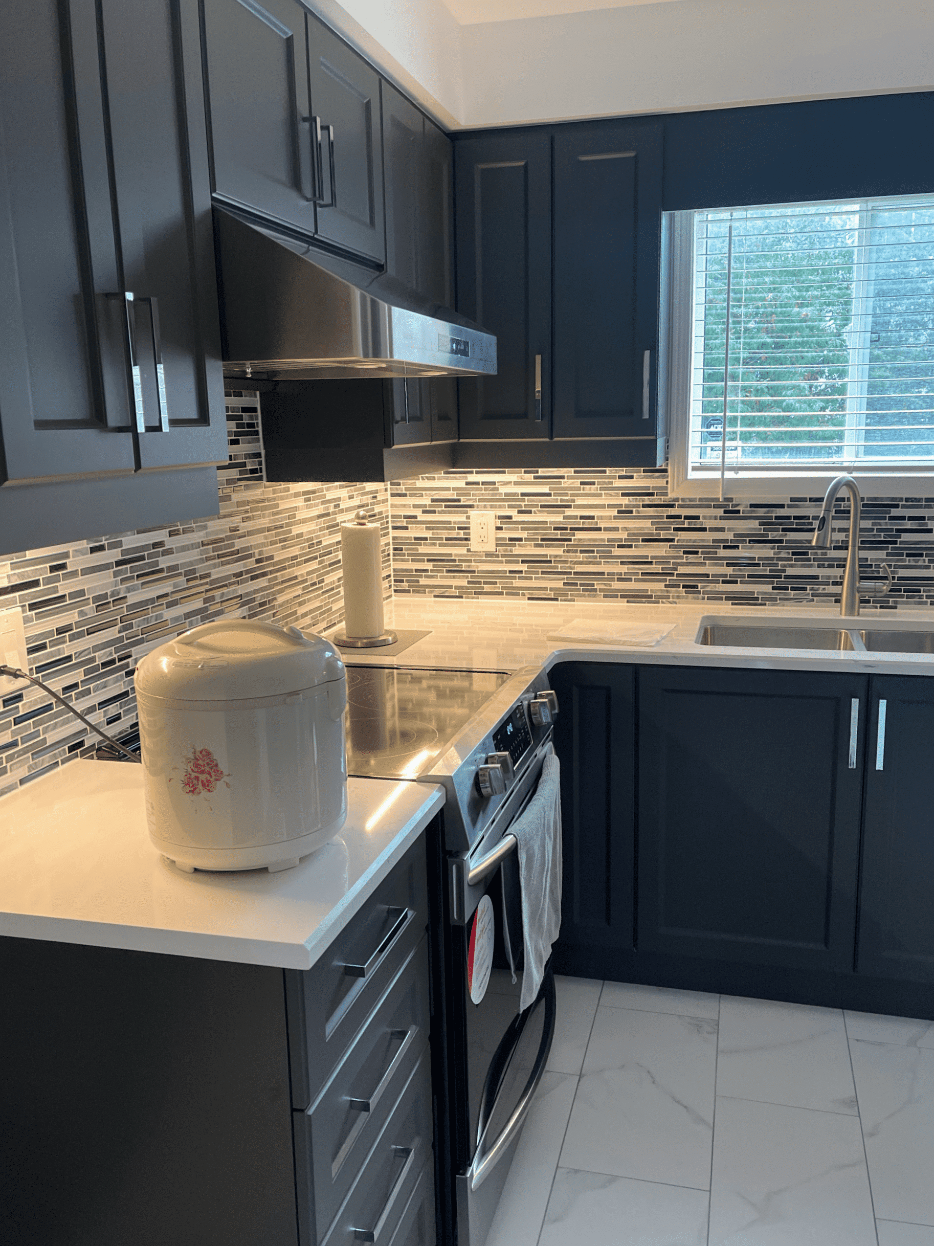 Modern kitchen with dark cabinetry, stainless steel appliances, and a stylish backsplash, featuring a countertop rice cooker and a bright window.