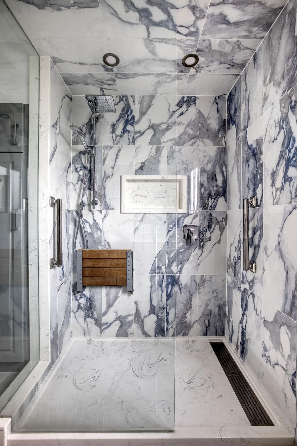 Modern bathroom shower featuring elegant gray and white marble tiles, overhead showerhead, and wooden fold-down seat, showcasing Superior Bath's custom renovation craftsmanship.