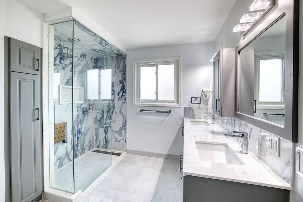 Modern bathroom renovation featuring a glass shower with marble walls, dual sinks with gray cabinetry, and natural light from a window, showcasing Superior Bath's custom design capabilities.