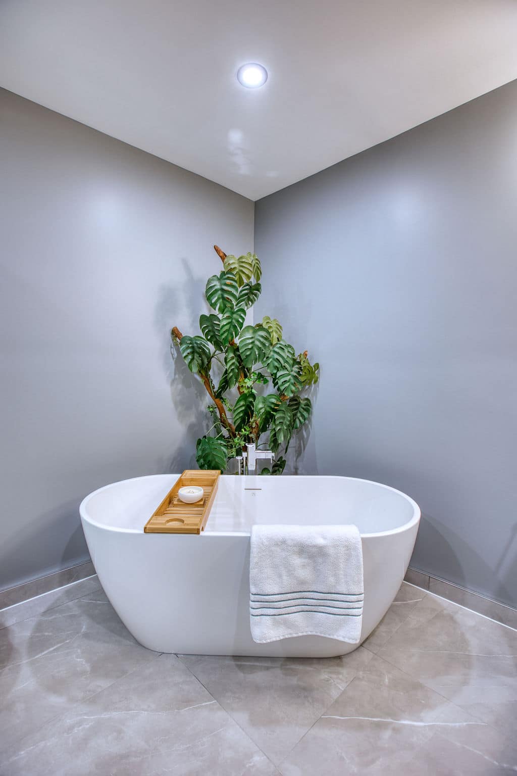 Modern custom bathroom featuring a freestanding white bathtub with a wooden bath tray, surrounded by green plants and grey walls, showcasing Superior Bath's design capabilities.
