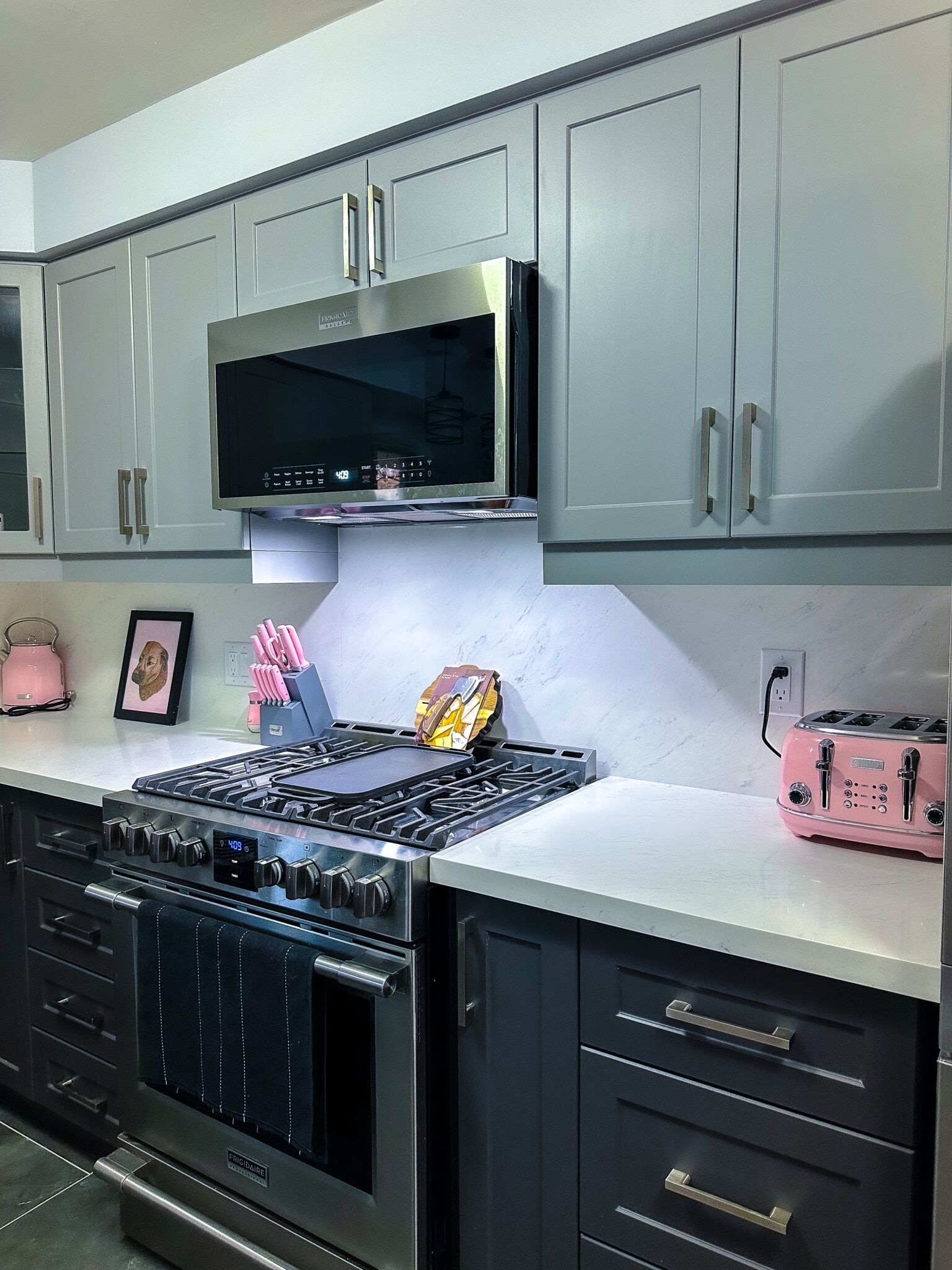 Modern kitchen featuring gray cabinetry, stainless steel appliances, a gas range with a black towel, pink kitchen accessories, and white marble countertops, showcasing Superior Bath's renovation style.