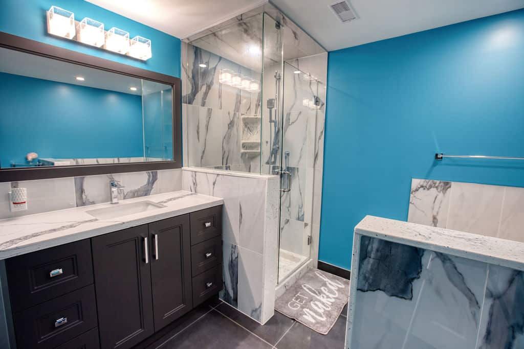 Custom bathroom featuring modern design elements, blue accent wall, marble countertops, sleek cabinetry, glass shower enclosure, and decorative bath mat with "GET NAKED" text.