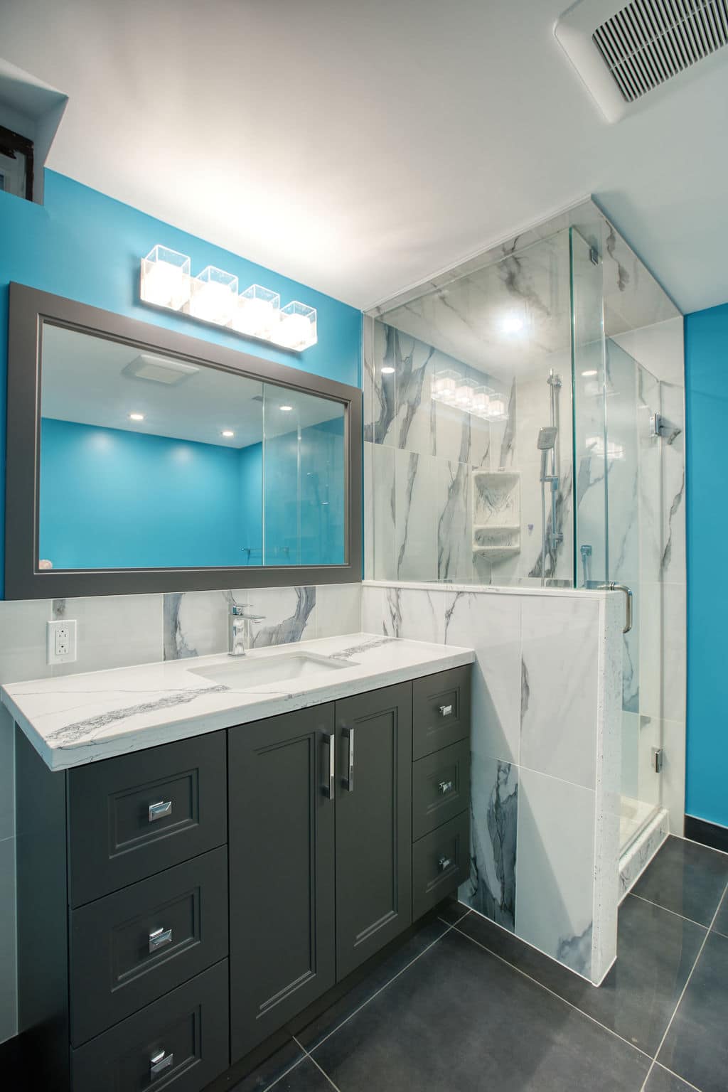 Custom bathroom design featuring a dark cabinetry with a marble countertop, illuminated mirror, and a glass shower enclosure against a vibrant blue wall, showcasing Superior Bath's renovation craftsmanship.